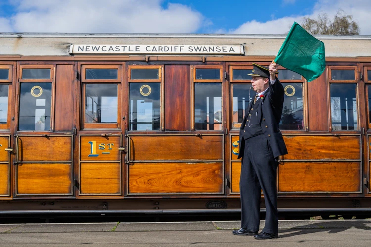 A conductor signals beside a vintage train carriage