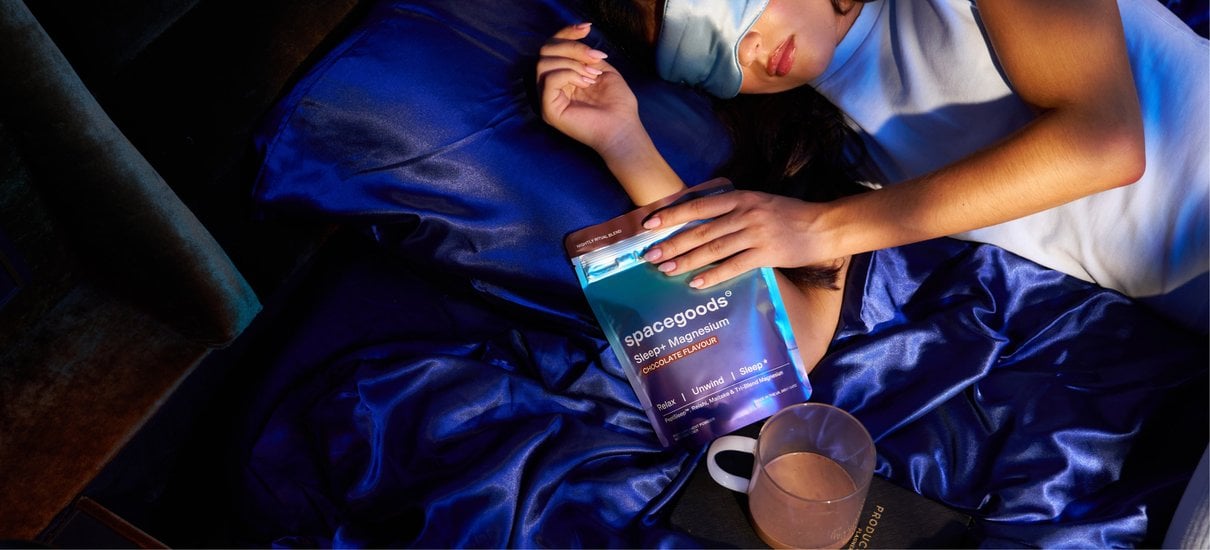 Person relaxing in bed with a mug beside Sleep+ Magnesium